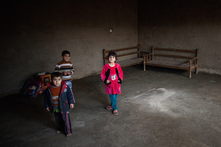 Three displaced children in Baghdad stand in their temporary home. The space is dark and empty, the concrete walls and floor bare.