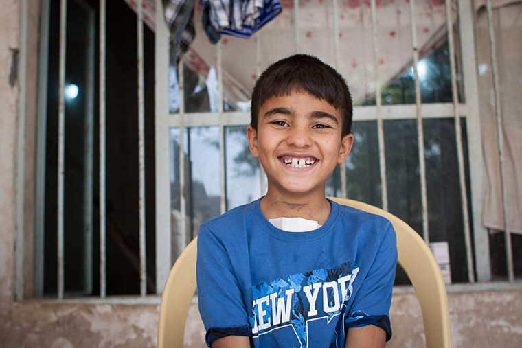 Mohammad sits in a chair in front of his house, beaming a huge smile. A fresh bandage peeks out of the top of his shirt. Mohammad recently had lifesaving heart surgery via Preemptive Love Coalition.